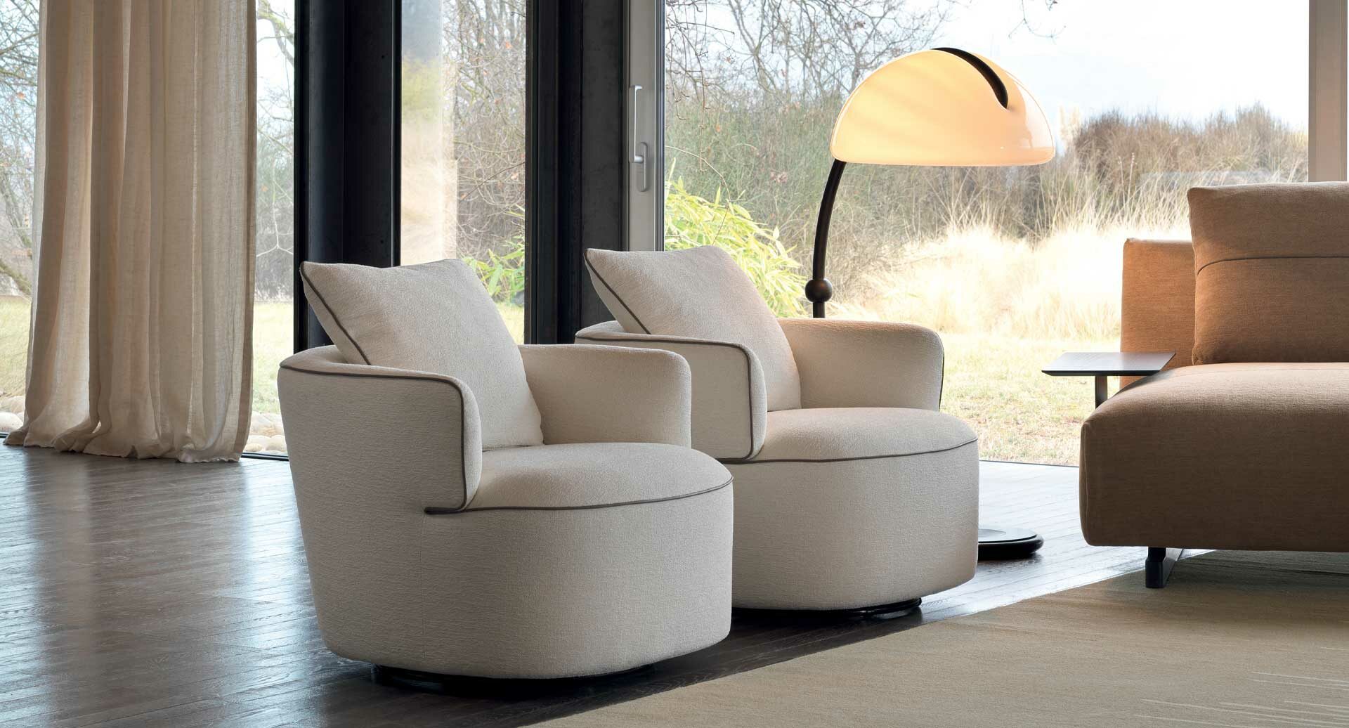 Modern living room with beige armchairs and a lamp near large windows.